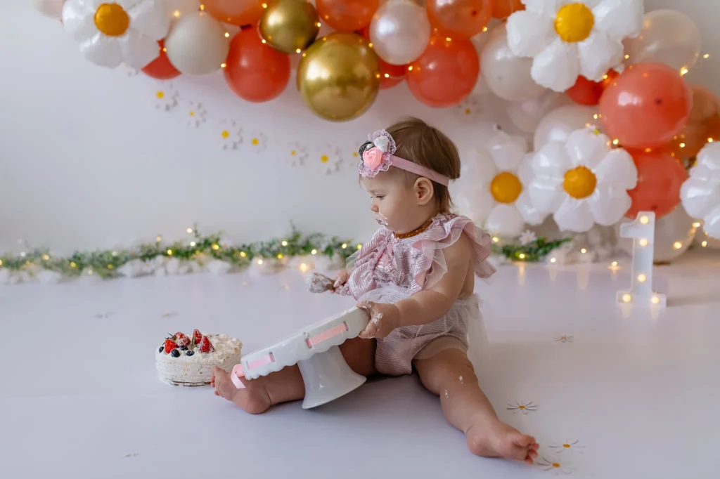 Cake smash fotosesija pirmo gimtadienio proga, vaikas sėdi šale torto ir rankoje turi torto gabalėlį